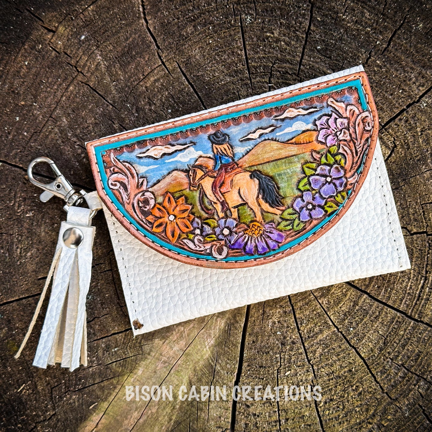 Handmade tooled leather cowgirl wallet with a western horse-riding scene, floral carving, and white leather body, displayed on rustic tree trunk background.