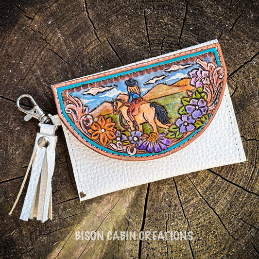 Handmade tooled leather cowgirl wallet with a western horse-riding scene, floral carving, and white leather body, displayed on rustic tree trunk background.