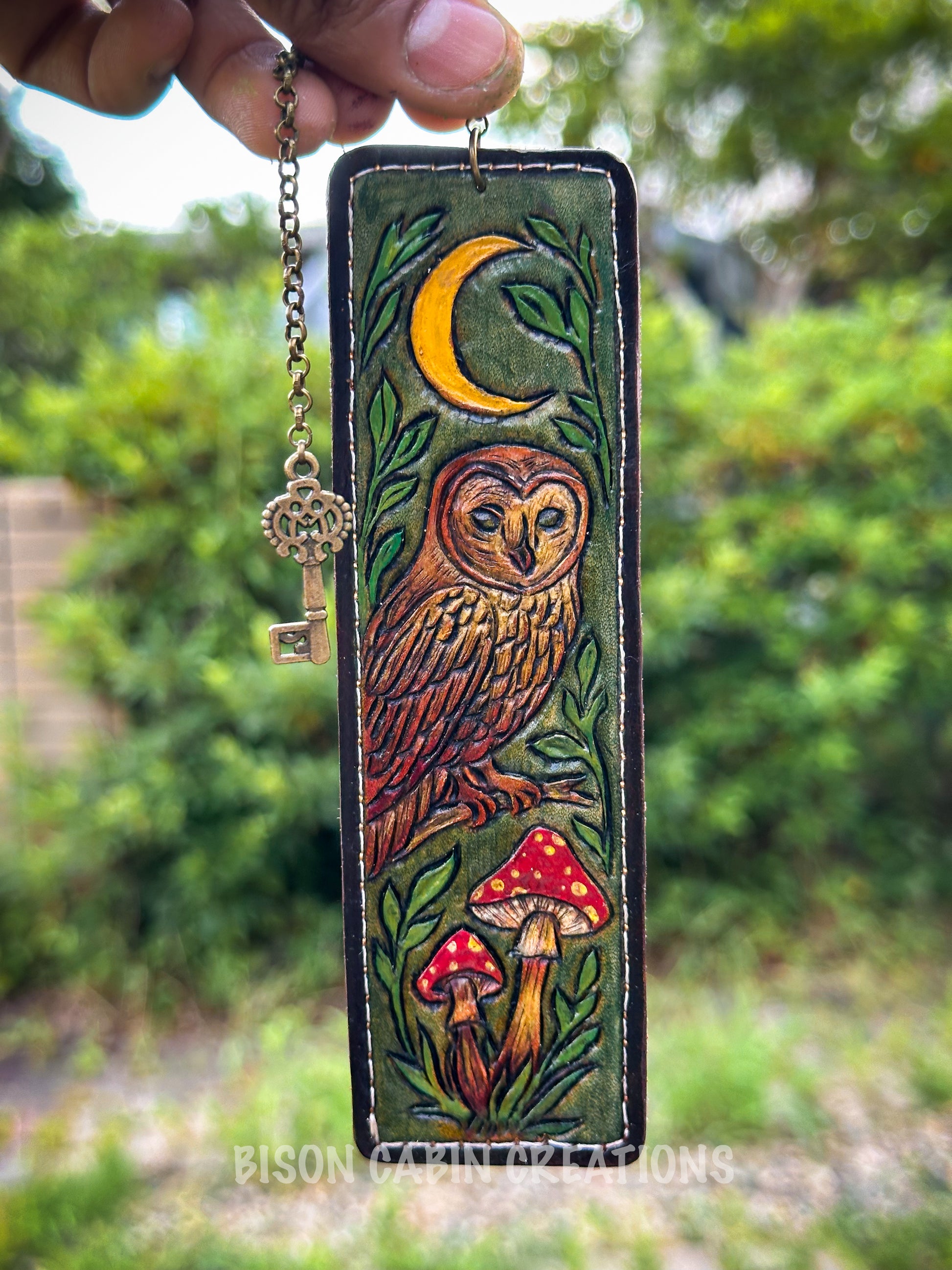 Decorative keychain with owl and mushroom design held in front of a blurred green outdoor background. Hand-tooled genuine leather bookmark featuring an intricate owl design, inspired by woodland nature