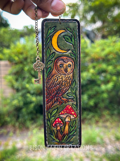 Decorative keychain with owl and mushroom design held in front of a blurred green outdoor background. Hand-tooled genuine leather bookmark featuring an intricate owl design, inspired by woodland nature