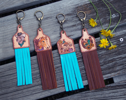 Collection of handcrafted, tooled leather keychains featuring horse, cow, dog, and horseshoe designs with rustic Western detailing and charm accessories